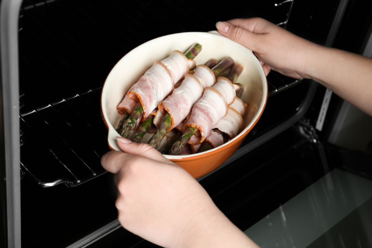 女人把装有熏肉包裹芦笋的陶瓷烤盘放进烤箱，特写